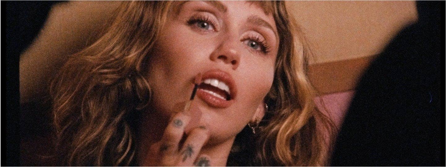 Miley Cyrus mit langen, gewellten Haaren posiert in einem Aufnahmestudio, bekleidet mit einem schwarzen Korsett-Oberteil und Hosen, Tattoos auf ihren Armen.