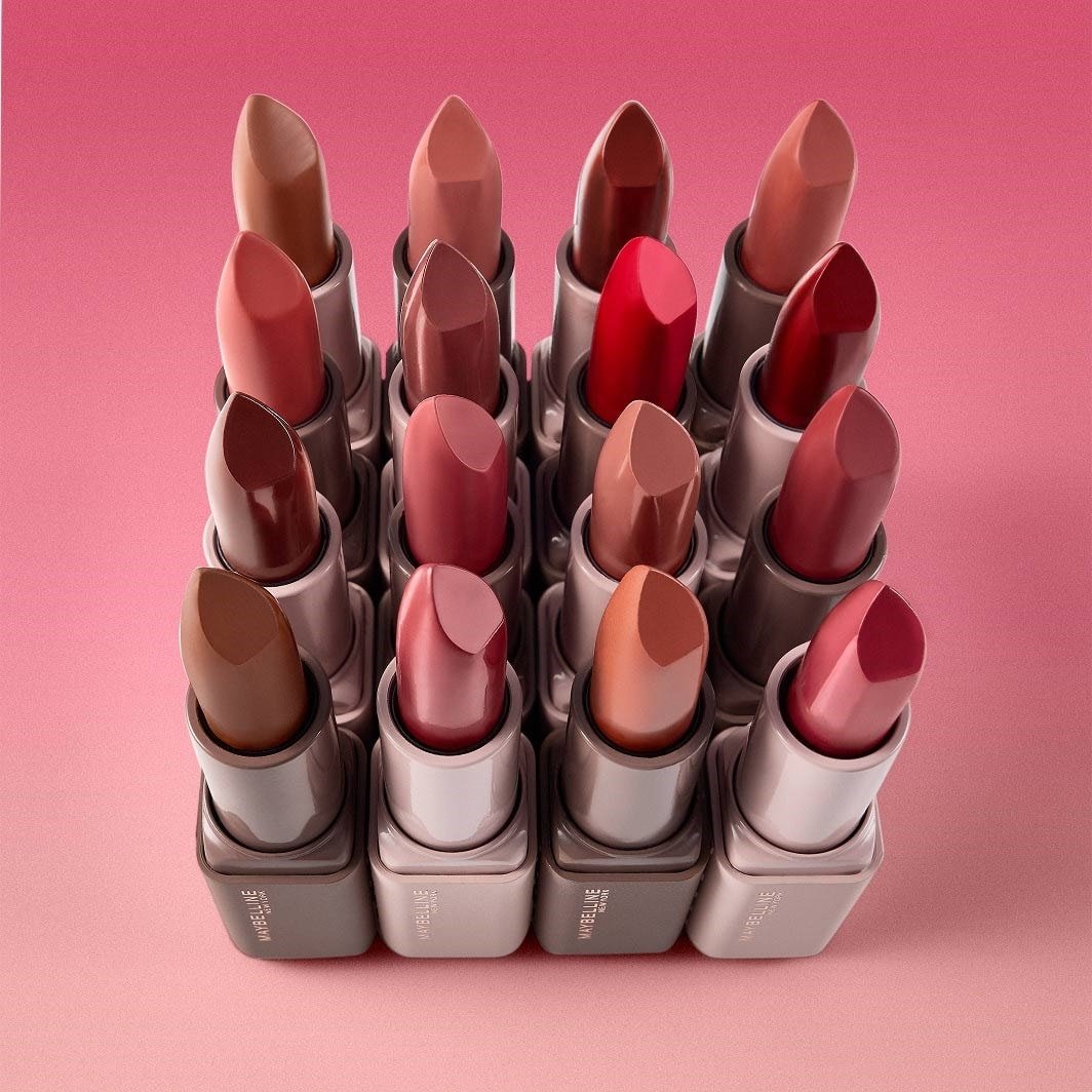Gros plan sur une grille de rouges à lèvres Maybelline dans différentes nuances de nude, rose, rouge et marron, disposés sur un arrière-plan rose.